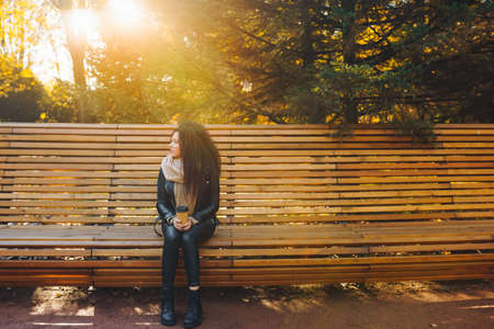 Sad young afro-haired woman sit alone on a wooden bench in autumn park at sunny warm day. Portrait of alone woman outdoor.の写真素材