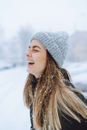 Outdoor portrait of young woman in warm jacket and knitted hat, turning her face to falling...