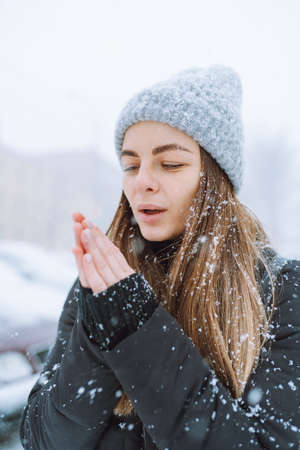 Outdoor portrait of young woman in warm jacket and knitted hat, turning her face to falling snowflakes.の写真素材