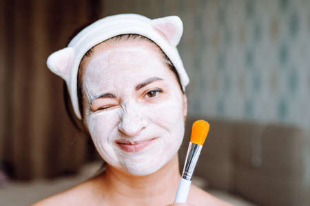 Woman with a white clay mask on her face holds a brush. Female takes care of the skin at home during...