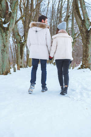 Young happy couple smile to each other and walk in winter park holding hands. Man and woman have fun together during winter holidays.の写真素材