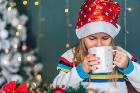 Little smiling girl Santa helper drink hot chocolate with marshmallows from christmas mug with with christmas wreath on backgroundの写真素材