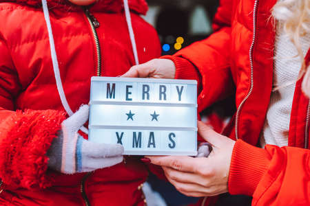Cropped image of mother and daughter wearig red lackets holding lightbox with inscription Merry Christmasの写真素材