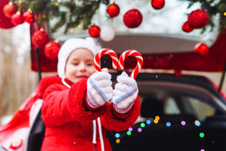 Blonde girl in red jacket and white hat have fun with red and white sweet sticks in decorated christmas car.の写真素材