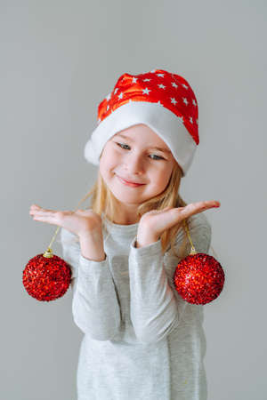 Cute blonde toddler girl wearing a santa hat holding two red Christmas balls of Christmas tree decoration against neutral background and smilingの写真素材