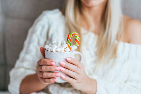 Blonde woman wearing white knitted dress hold mug with dhot cocoa with marshmallows and candy cane. Christmas mood.の写真素材
