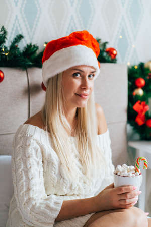 Blonde woman wearing white knitted dress Santa hat sit on bed in Christmas-decorated bedroom and drink hot cocoa with marshmallows.の写真素材
