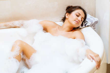 Young woman lies in white bath of foam with her eyes closed. Spa procedures, cleansing, relaxation, self-care.の写真素材