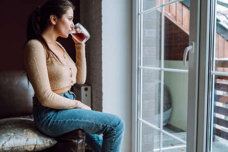 Sad young woman look out the large panoramic window from home, holding cup of tea. Loneliness,...