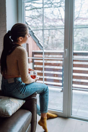 Sad young woman look out the large panoramic window from home, holding cup of tea. Loneliness, depression, divorce, melancholy.の写真素材