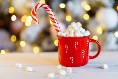 Red mug of hot chocolate with marshmallow and candy stick against the background of the lights of...