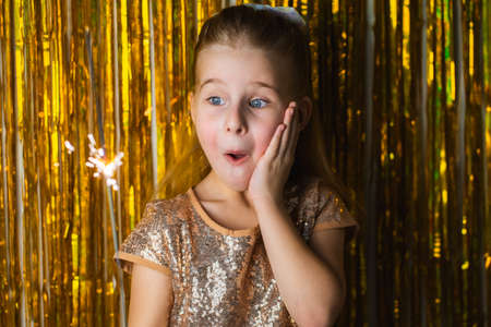 Cute blonde toddler girl wearing fancy hold burning sparkler against golden glitter background and...