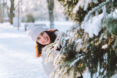 Young curly smiling woman walk in winter park and hide behind snow-covered Christmas tree. Positive emotions, freshness, holidaysの写真素材