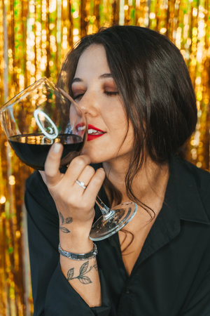 Smiling young woman with long hair, red lips hold wine glass touching with lips wearing black suit on tinsel background.の写真素材