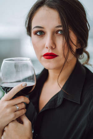 Portrait of serious young woman with long black hair, red lips hold wine glass wearing black suit on urban background.の写真素材