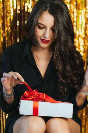 Young woman with long hair and red lips pull ribbon looking at present wearing black suit on golden tinsel background.の写真素材