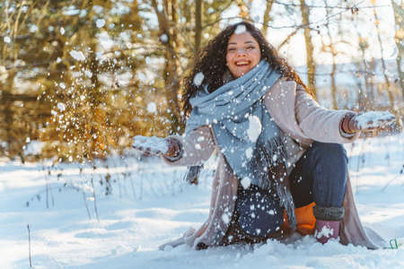 Smiling beautiful young woman have fun throwing up snow wearing warm clothes and blue stole in snowy winter forest.の写真素材