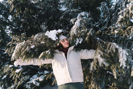 Young curly smiling woman walk in winter park and hide behind snow-covered Christmas tree. Positive emotions, freshness, holidaysの写真素材