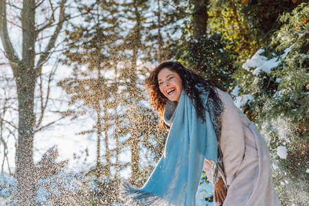Happy laughing young woman have fun under falling snow wearing warm clothes and blue stole in snowy...