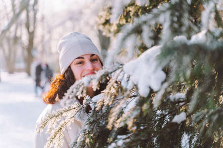 Young curly smiling woman walk in winter park and hide behind snow-covered Christmas tree. Positive emotions, freshness, holidaysの写真素材