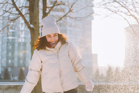 Smiling curly young woman have fun playing with snow wearing warm white clothes in snowy winter...