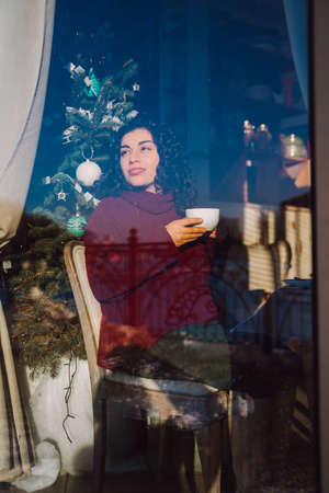 Young happy smiling curly woman drink tea in city cafe and look out the window with decorated...