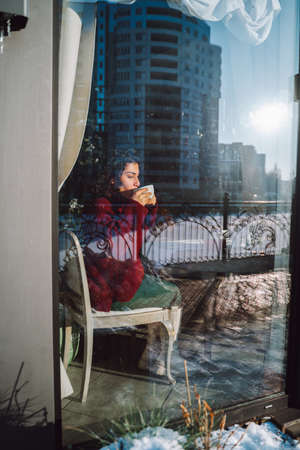 Young happy smiling curly woman drink tea in city cafe and look out the window with decorated Christmas tree in background.の写真素材