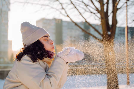 Side view of young positive woman blowing on the snow in her palms in winter park on cold sunny dayの写真素材