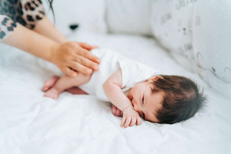 Cute little newborn baby with dark hair in white bodysuit lie sidelong smiling and mother touching...