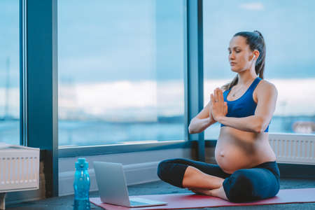 Meditation during pregnancy. Pregnant woman with closed eyes and her hands in namaste gesture sit on mat with laptop in lotus pose and practicing yoga online. Expectation, mindfulness, future motherの写真素材