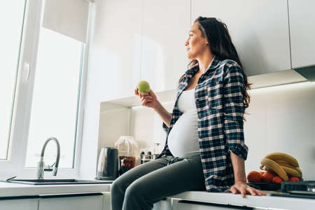 Eat healthy during pregnancy. Young pregnant woman in casual outfit sitting on table in light modern...