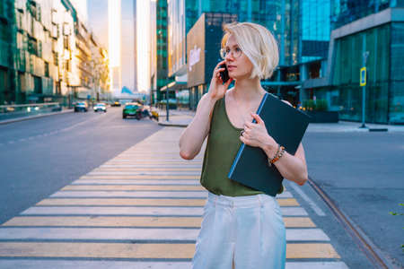 Young blonde businesswoman with laptop making call by phone outdoors against skyscrapers. Business...