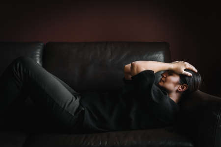 Unhappy woman dressed in black lying on black sofa keeping her hands on her head experiencing negative emotions possibly depression, grief, stress, loneliness, loss, fear, dissipation, sacrificeの写真素材