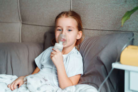 Sick little girl making inhalation with nebulizer to reduce coughing, lying in bed at home, child...
