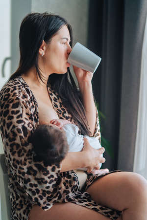 Young long dark-haired woman mother in leopard print pattern bathrobe breastfeeding holding baby in arms. Female sit and drink coffee near window in sunny light day , side viewの写真素材