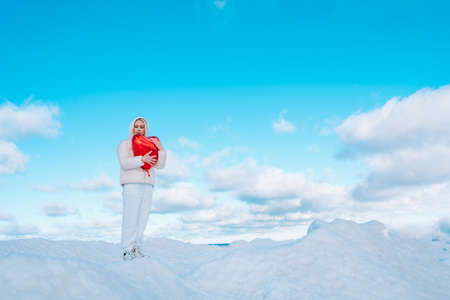 Portrait of young woman in white clothes with red heart balloon waiting for love on holiday blue cloudy sky on background. Sky merges with earth near snowdrifts. Side view. Copy space. Minimalismの写真素材
