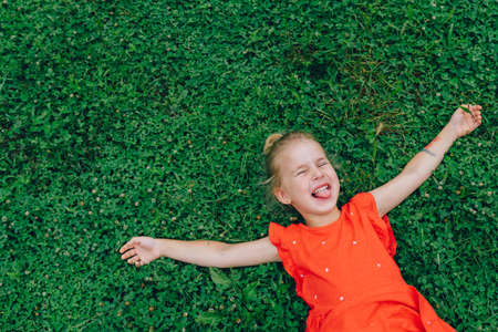 Portrait of little girl in red dress lying on soft grass with arms spread and eyes closed tight.Positive and laughing concept. Top view. Green plants leaf background.Tongue hanging out. Copy space.の写真素材