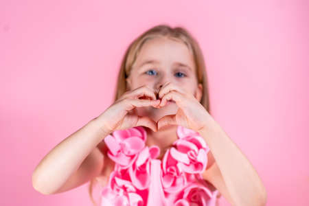 Portrait of beautiful little blue-eyed girl with long fair hair in chic pink dress pose showing heart shape with hands on pink background. Valentines day, love, friendship, family, emotion concept.の写真素材