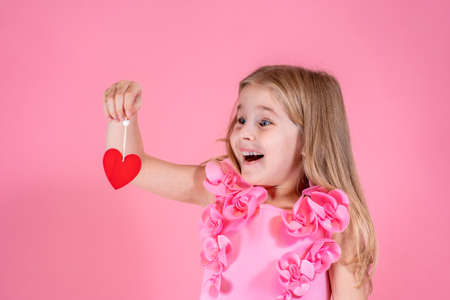 Funny little blue-eyed girl with long fair hair in chic pink dress stand smiling looking at red paper heart on string in amazement on pink background. Valentines day, love, emotion concept.の写真素材