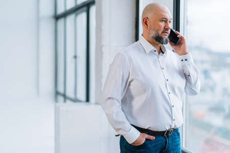Serious mature businessman in white shirt and jeans talking by phone with client or partner while standing near panoramic window in office, selective focus. Mobile technology in business communicationの写真素材
