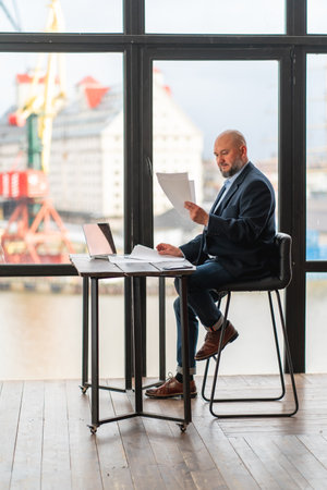 Full-length of focused mature businessman analyzing documents, doing paperwork while sitting at office desk with laptop near panoramic window with construction site view. Work at midlifeの写真素材