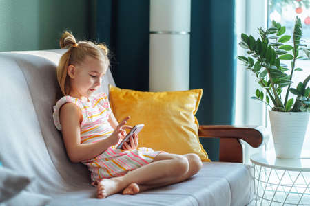 Little girl surfing wireless internet on smartphone, application messenger, sitting on sofa in living room. Covid coronavirus isolation, lockdown. Social chatting, remote distance communication. Sideの写真素材
