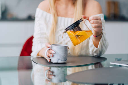 Unrecognisable woman in kitchen pouring drinking yellow lemon orange herb tea teapot. Wearing knitting white sweater, bracelet, sitting glass table, red soft chair. Blurred kitchen-ware backgroundの写真素材