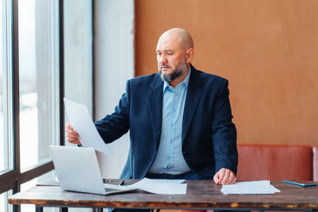 Bald adult businessman beard working doing paperwork, in suit jacket, sitting wooden desk laptop, reading contract, deal, smartphone, light office window. Thinking strategy plans, start up marketingの写真素材
