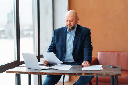Bald adult businessman beard working doing paperwork, in suit jacket, sitting wooden desk laptop, signing contract, deal, smartphone, light window. Light brown sofa back office. Not pleased agreementの写真素材