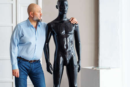Bald sad adult man with beard, holding shoulders of naked artificial plastic black manikin, looking to dummy, thinking of alopecia treatment. No hair, replacement medicine concept. Light indoorの写真素材