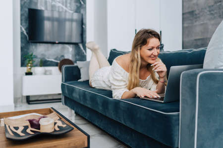 Blond attractive woman lying blue sofa pillow, surfing internet, shopping online relaxing weekend. Plate candle decor wooden table interior. Wearing white wool half-hose sweater dress.の写真素材