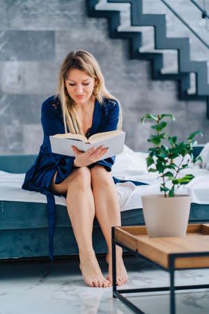 Vertical blond attractive barefoot smiling woman in sapphirine silk bathrobe reading thick book resting home on bed blue sofa near wooden table with flower pot plant. Stairs apartment on background.の写真素材