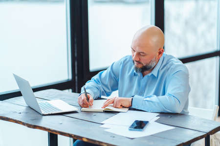Portrait of confident bald adult man with beard sitting table, smartphone,papers, laptop, window in office, signing contract, thinking about new investment. Wearing shirt, suit. Light office indoorの写真素材