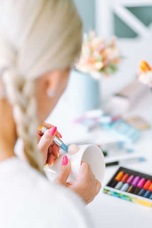 Vertical unrecognizable photo of woman artist hands with manicure jobbing and making decoration crafts flowers on mugs of polymer clay. Blurred color clay in box, cups with brushes, painters on tableの写真素材